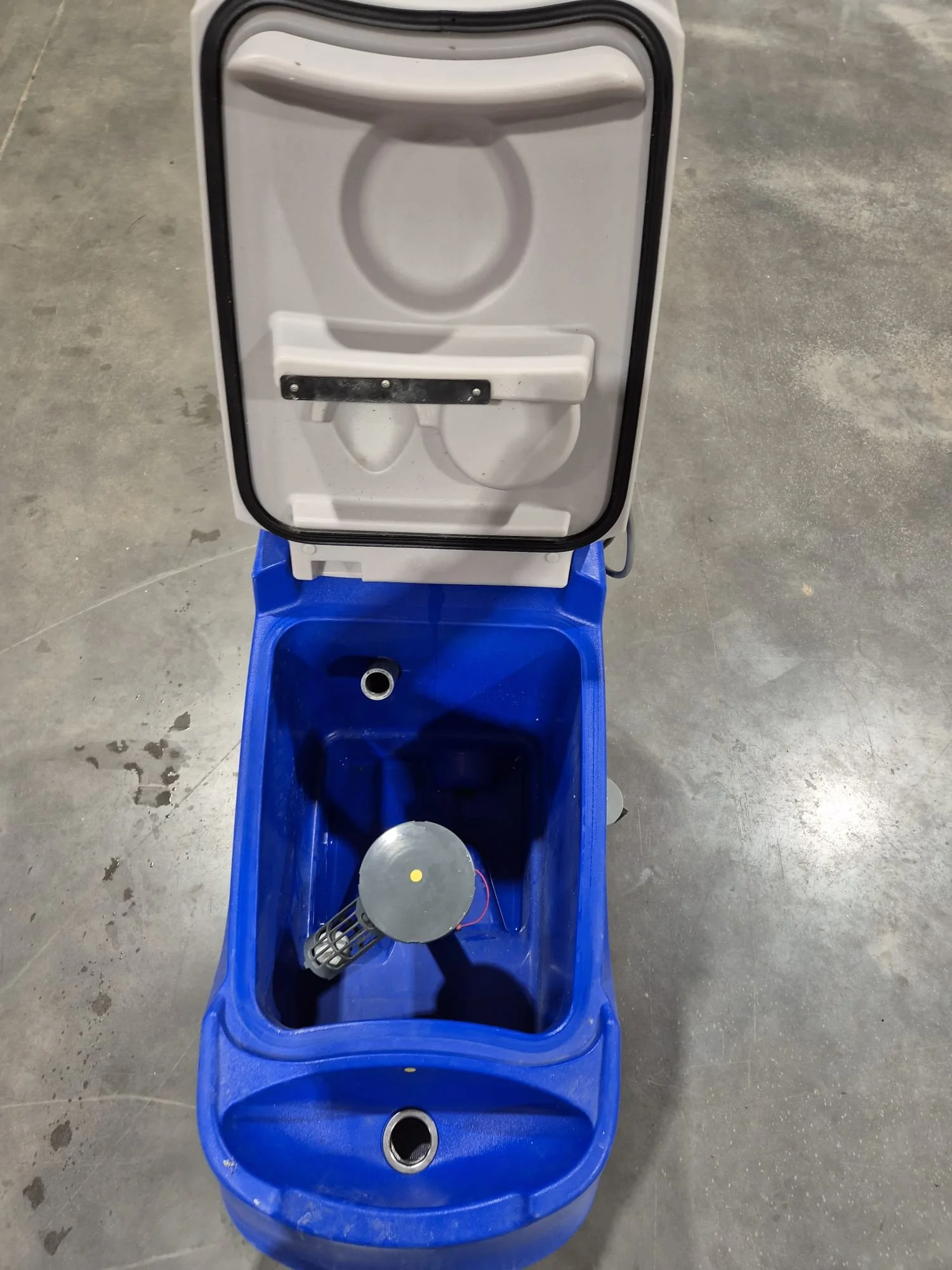 Ein blauer Toilettenwagen mit offenem Deckel, darin ein Wasserballonwerfer. Der Boden ist eine grau betonte Betonfläche.