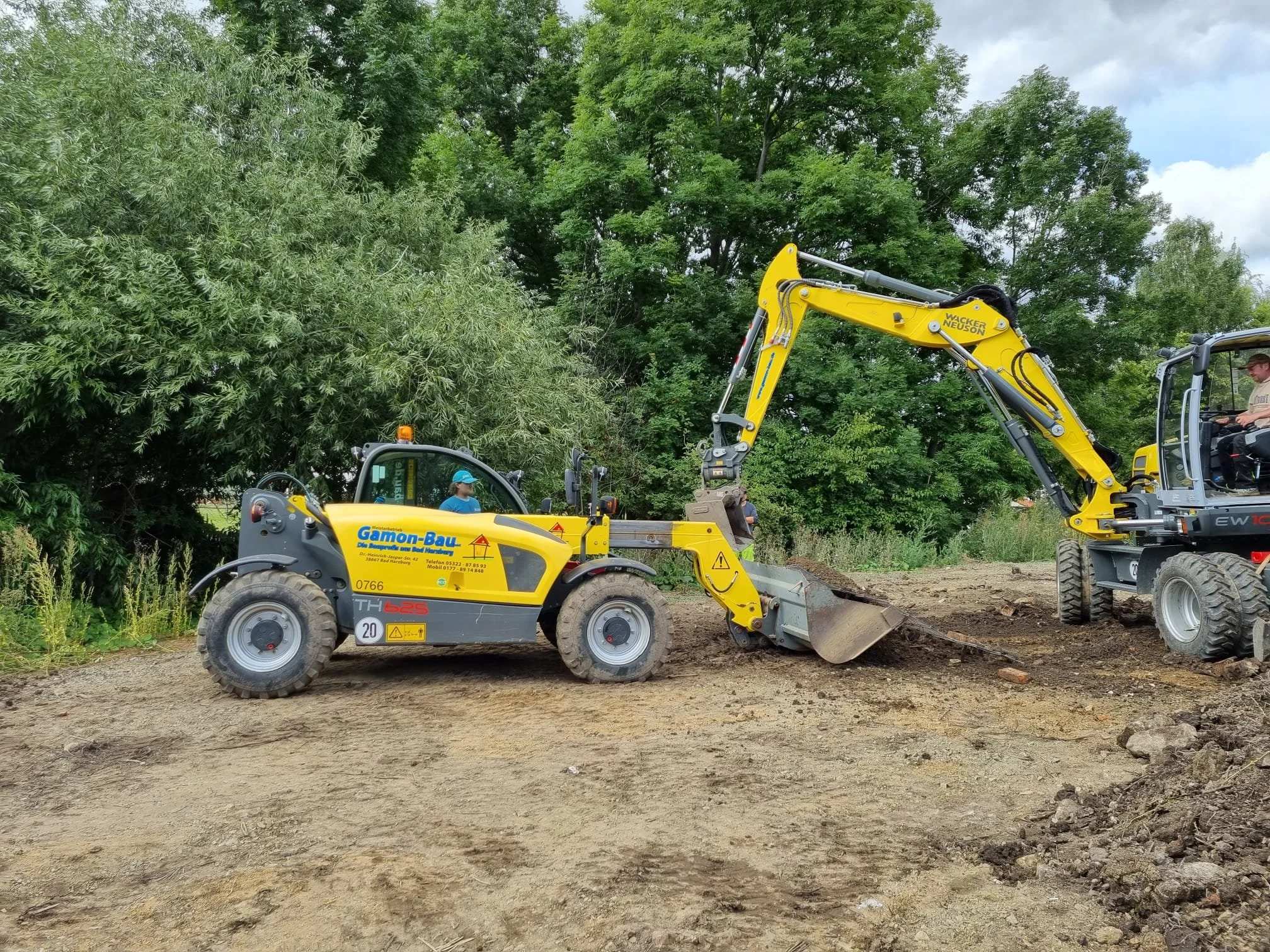 Zwei Baugeräte an einem Baugrundstück, eines ist ein Teleskoplader und das andere ein Mobilbagger. Die Baugeräte sind im Einsatz auf einer Baustelle im Freien in der Nähe von Bäumen und einem bewölkten Himmel.