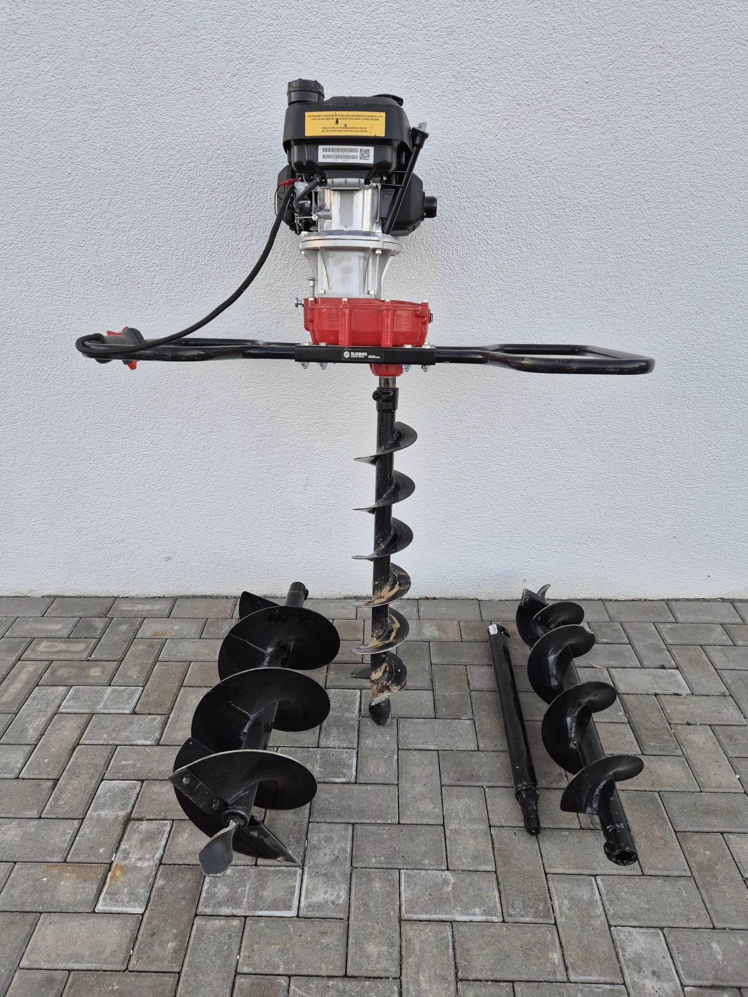 Ein Erdbohrer mit mehreren abnehmbaren Bohrern, der auf einer gepflasterten Fläche vor einer weißen Wand steht.