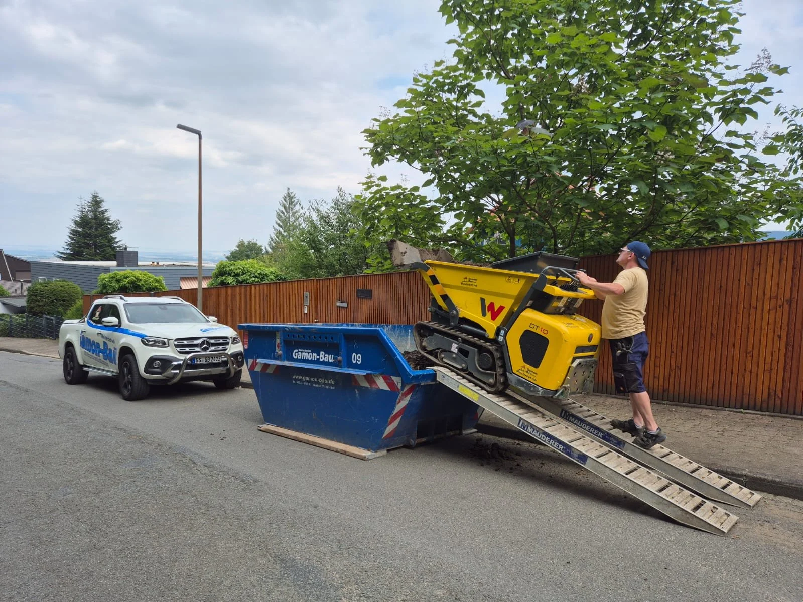 Mann steht auf einer Rampe und lädt einen kleinen, gelben Raupendumper in einen großen blaue Container. Im Hintergrund steht ein weißes Auto neben einem Holzzaun, umgeben von grünen Bäumen und einem blauen Himmel.