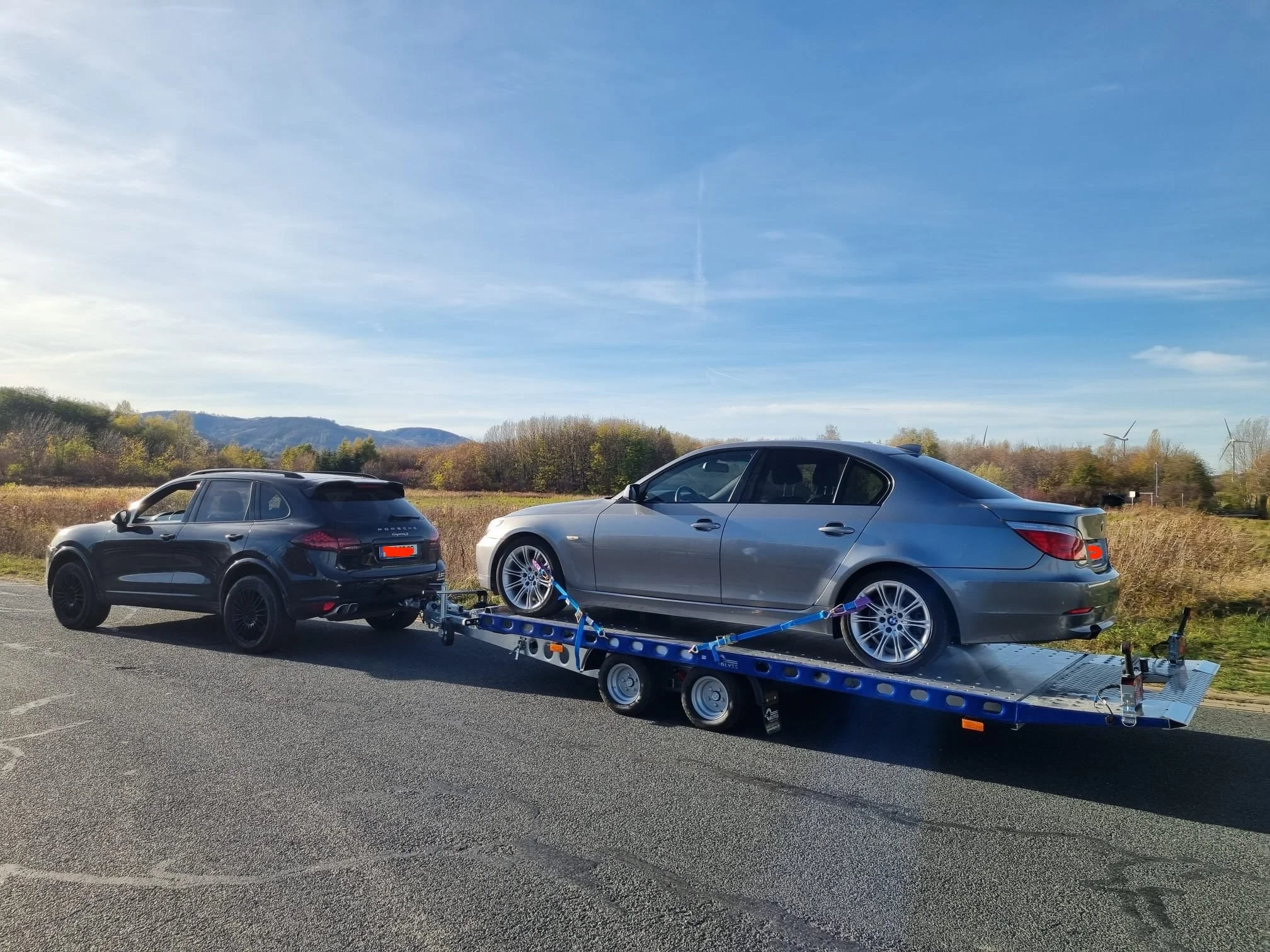 Ein Autotrailer mit einem schwarzen SUV und einem grauen Auto auf einer Landstraße bei sonnigem Wetter mit Himmel und Bäumen im Hintergrund.