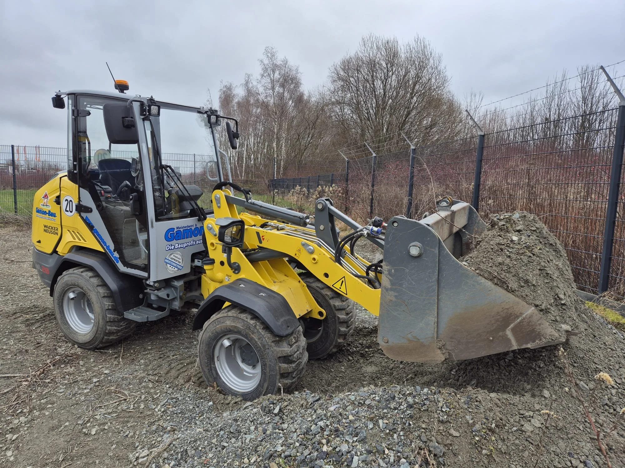 Radlader mit schwerem Baggerlöffel, der eine große Menge Erde oder Sand hebt, auf einer Baustelle hinter einem Zaun, bewölkter Himmel.