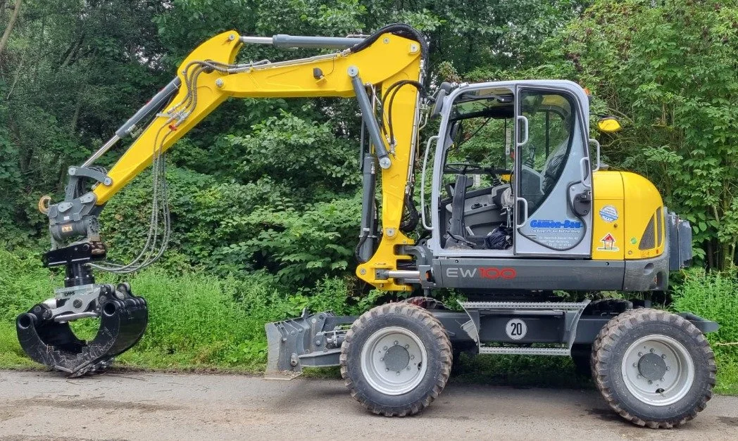 Gelber und grauer Kompaktbagger auf einer Straße, umgeben von Büschen und Bäumen, mit Schutzschilden und Anbauteilen.