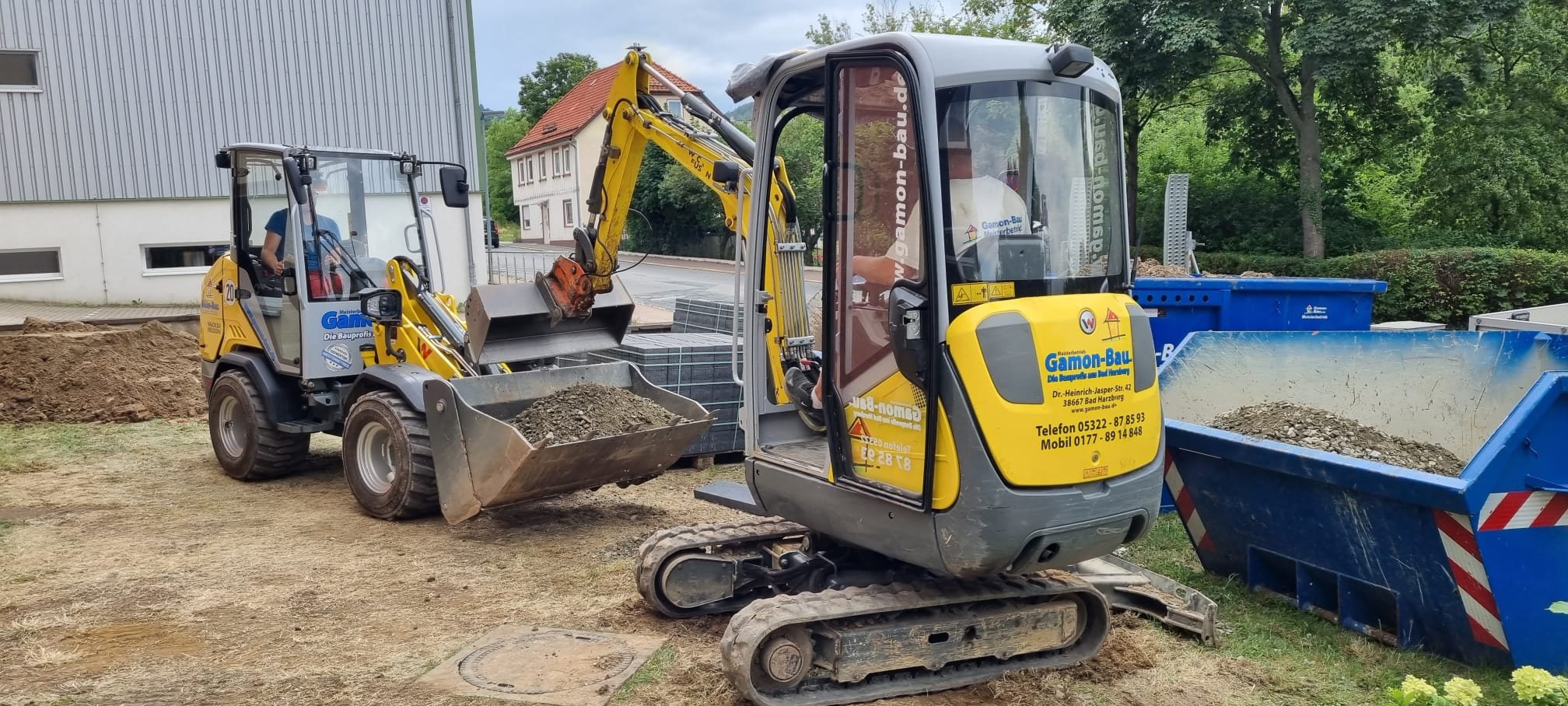 Kleiner Bagger- und Radlader auf einer Baustelle mit Schaufel Ladung Erde, im Hintergrund Bäume und Wohnhäuser.