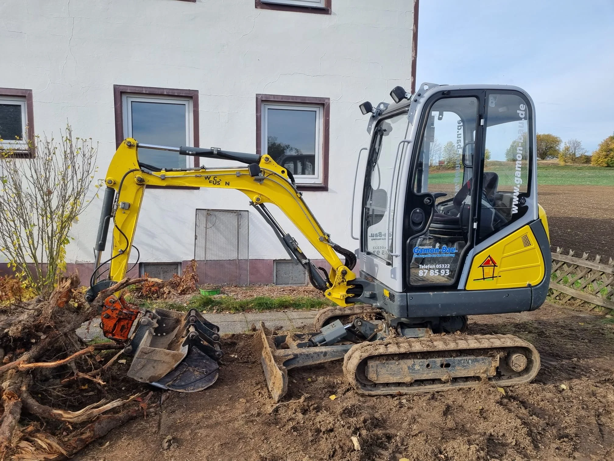 Kleine Baumaschine beim Graben am Haus, mit Gummibandketten und einem ausgestreckten Arm, stehend neben einem Garten. Im Hintergrund das Haus mit weißen Wänden und mehreren Fenstern, sowie ein Feld und Bäume.