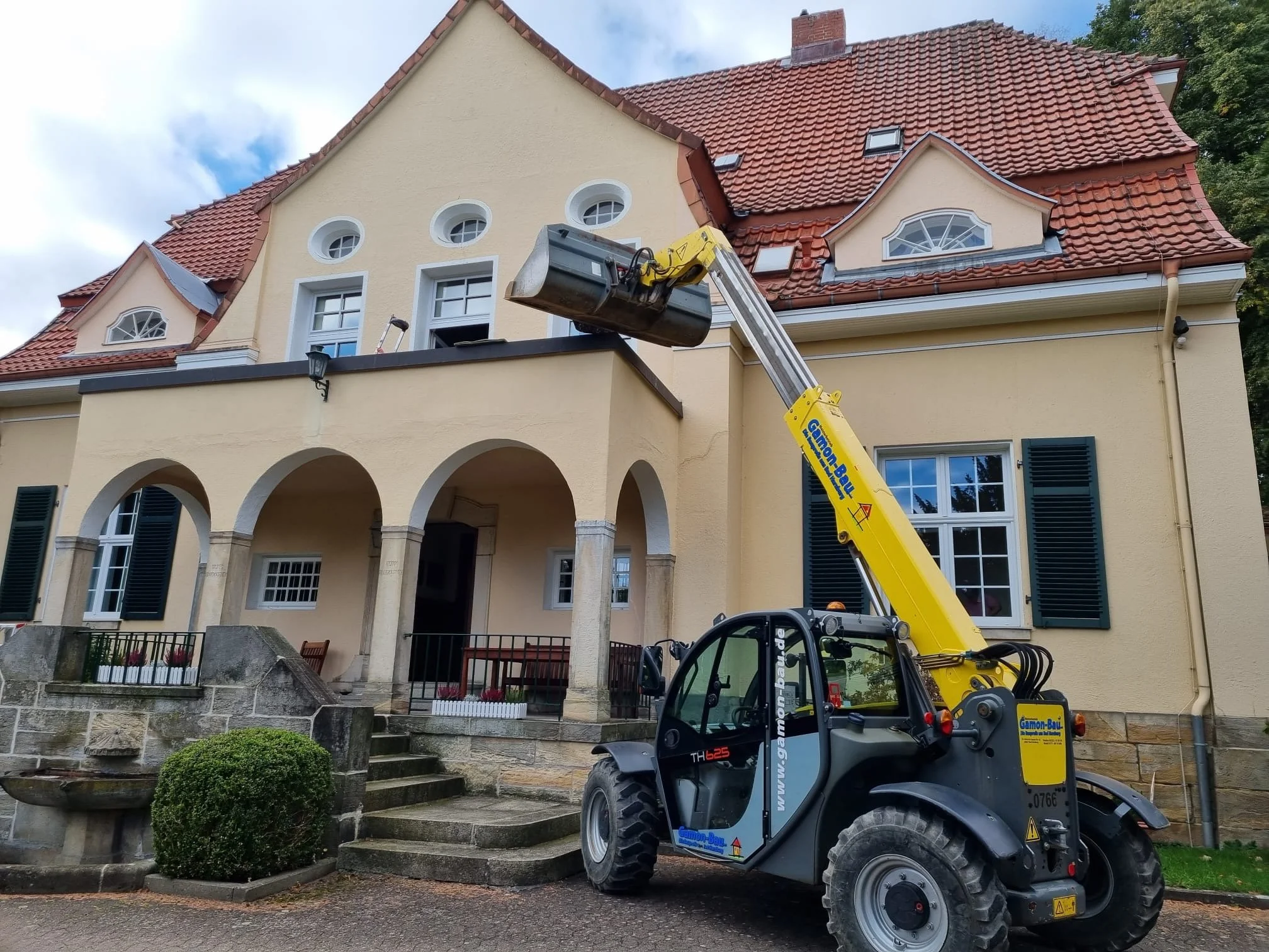 Ein Teleskoplader hält einen Lastenkorb vor einem mehrstöckigen Haus mit rotem Ziegeldach und gelben Wänden. Das Haus hat mehrere Fenster mit blauen Jalousien und eine Treppe führt zur Eingangstür.