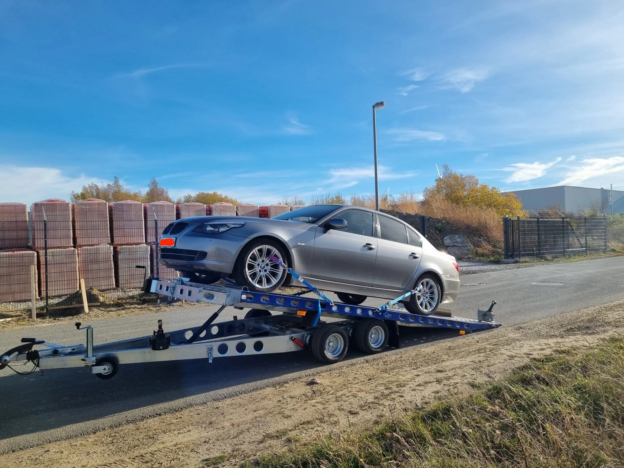 Ein silbernes Auto ist auf einem Abschleppwagen an einer Straße, im Hintergrund sind Bauzäune, Bäume und ein Gebäudekomplex bei blauem Himmel sichtbar.