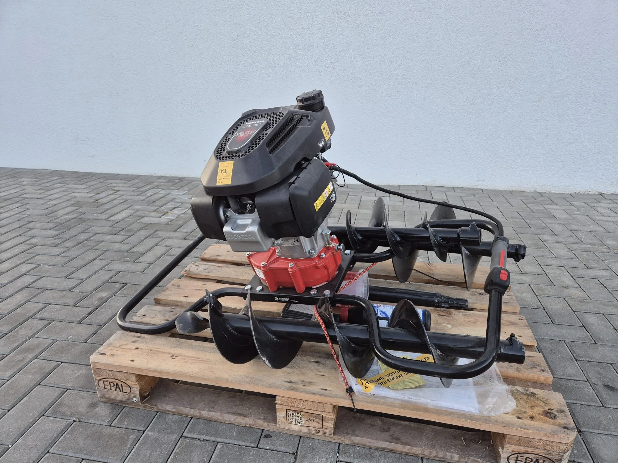 Ein motorisiertes Erdbohrgerät mit rot-schwarzem Motor und drei große Schrauben auf einem Holzpalette auf Pflasterstein