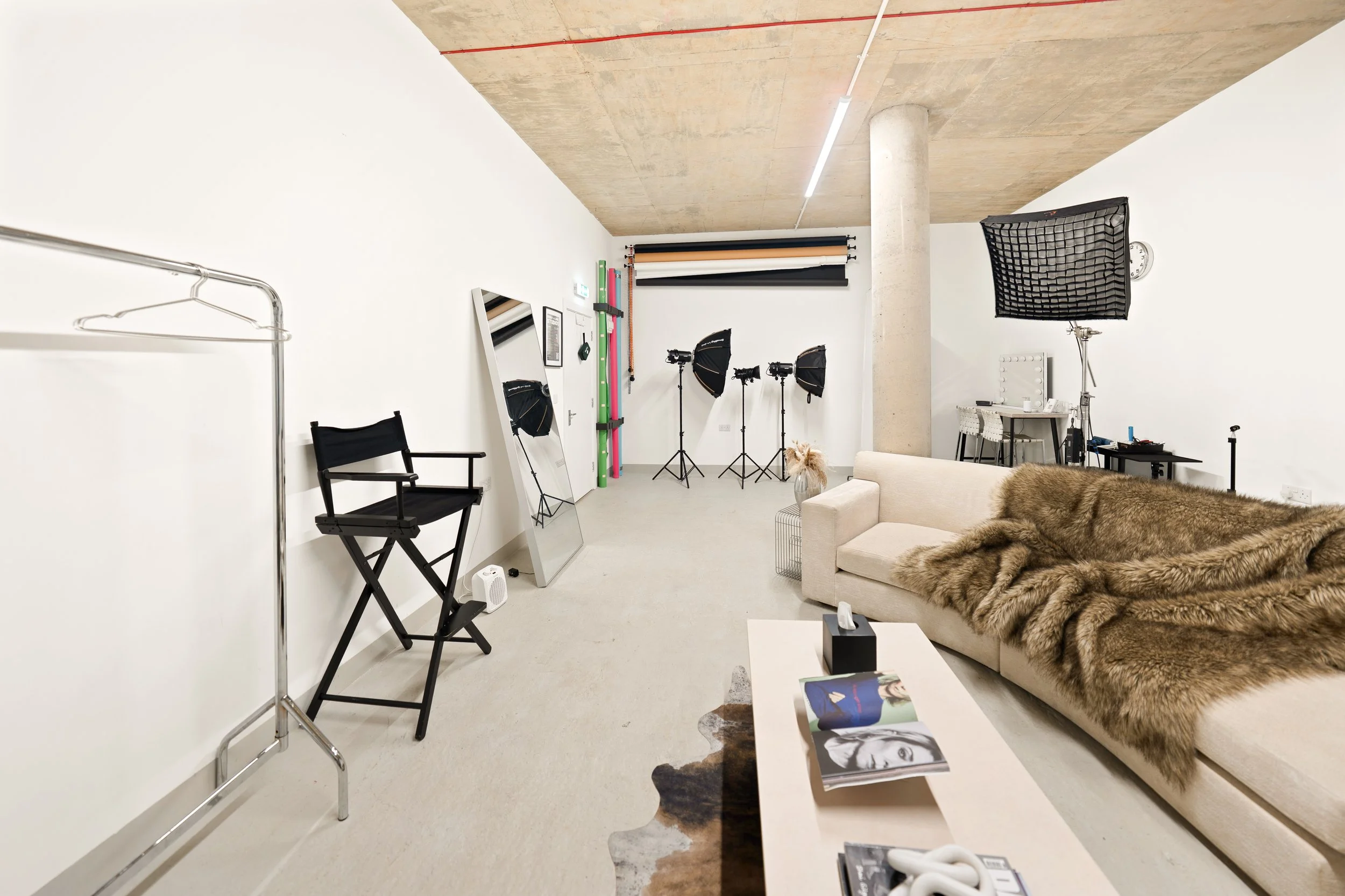 Photography studio setup with a white wall, a mirror, three studio lights, backdrop rolls, a director's chair, a sofa with a fur throw, and a coffee table with magazines.