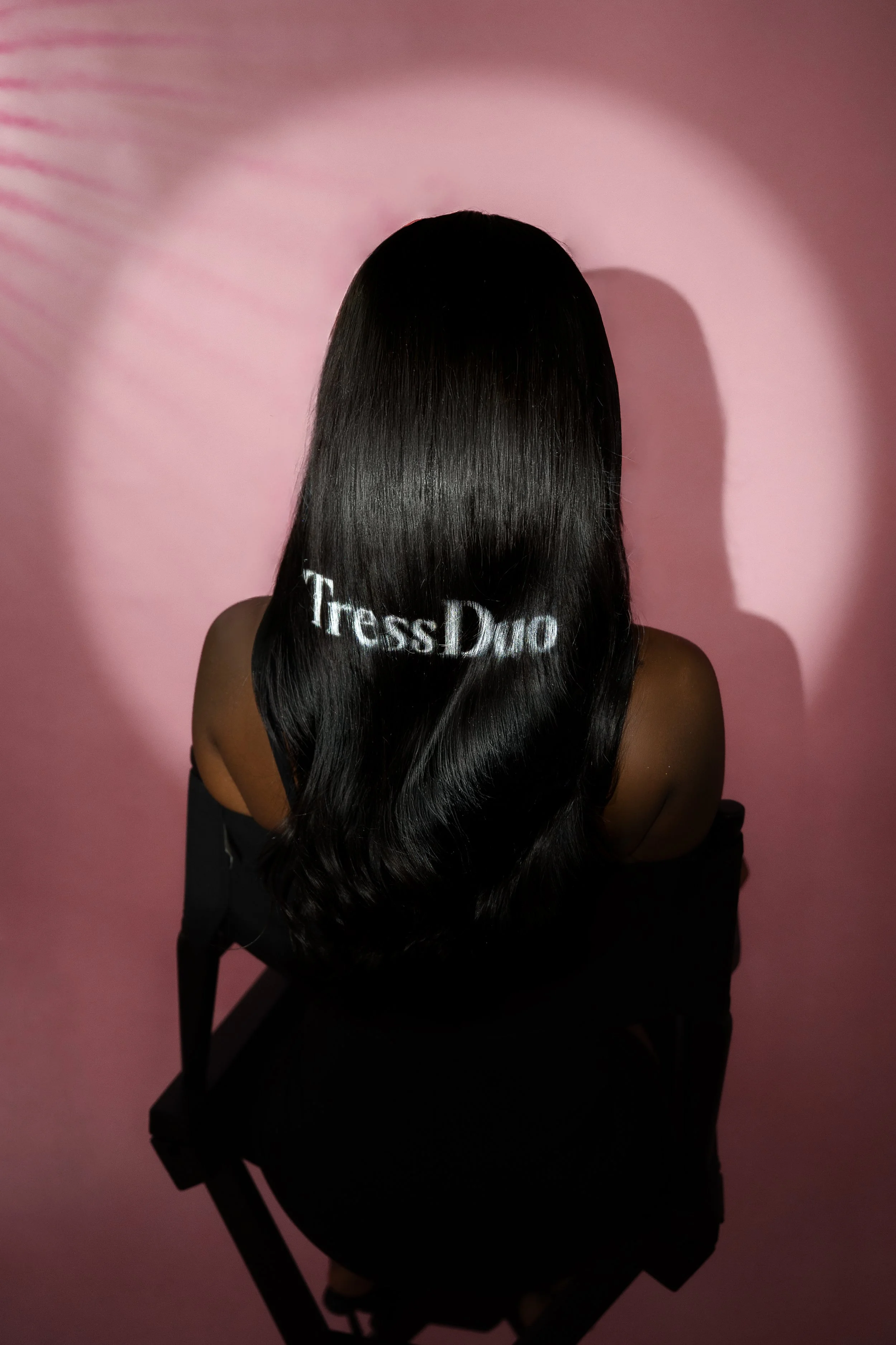A woman with long, black, shiny hair sitting in a black chair against a pink background, with a shadow on the wall. Creating content at a studio in East London for her wig business