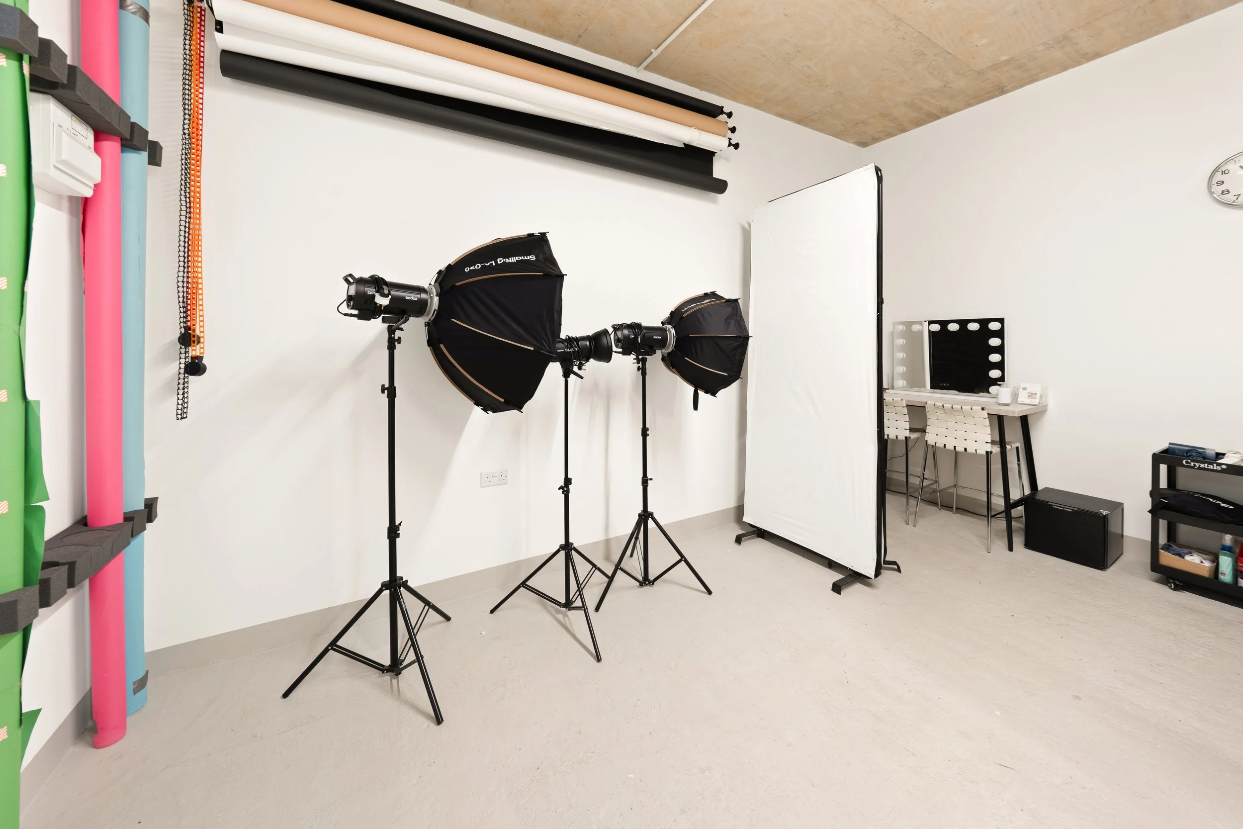 Photography studio with two softbox lights on stands, a white backdrop, a small table with makeup supplies, and fabric rolls on shelves along the wall.
