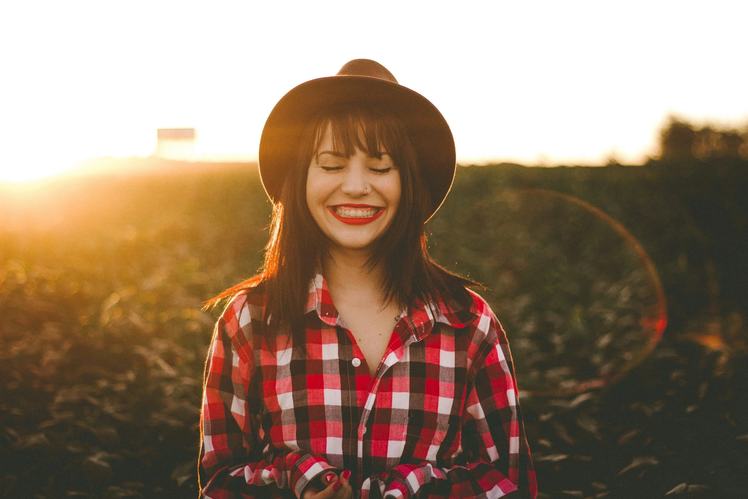 A young woman wearing a brown hat and red plaid shirt, smiling with her eyes closed, standing outdoors during sunset.