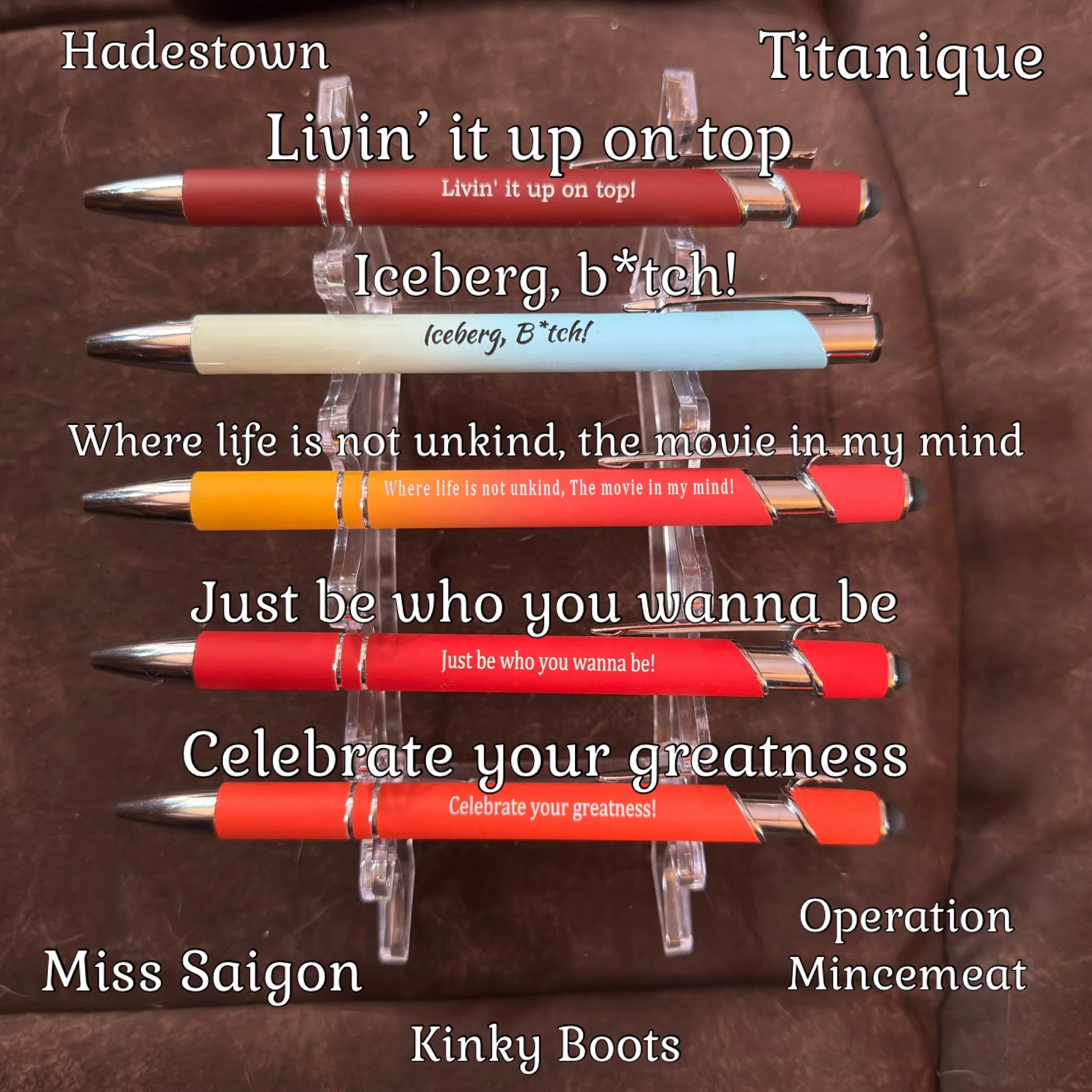 Five pens with various phrases and names written on them, displayed on a clear stand against a brown background.