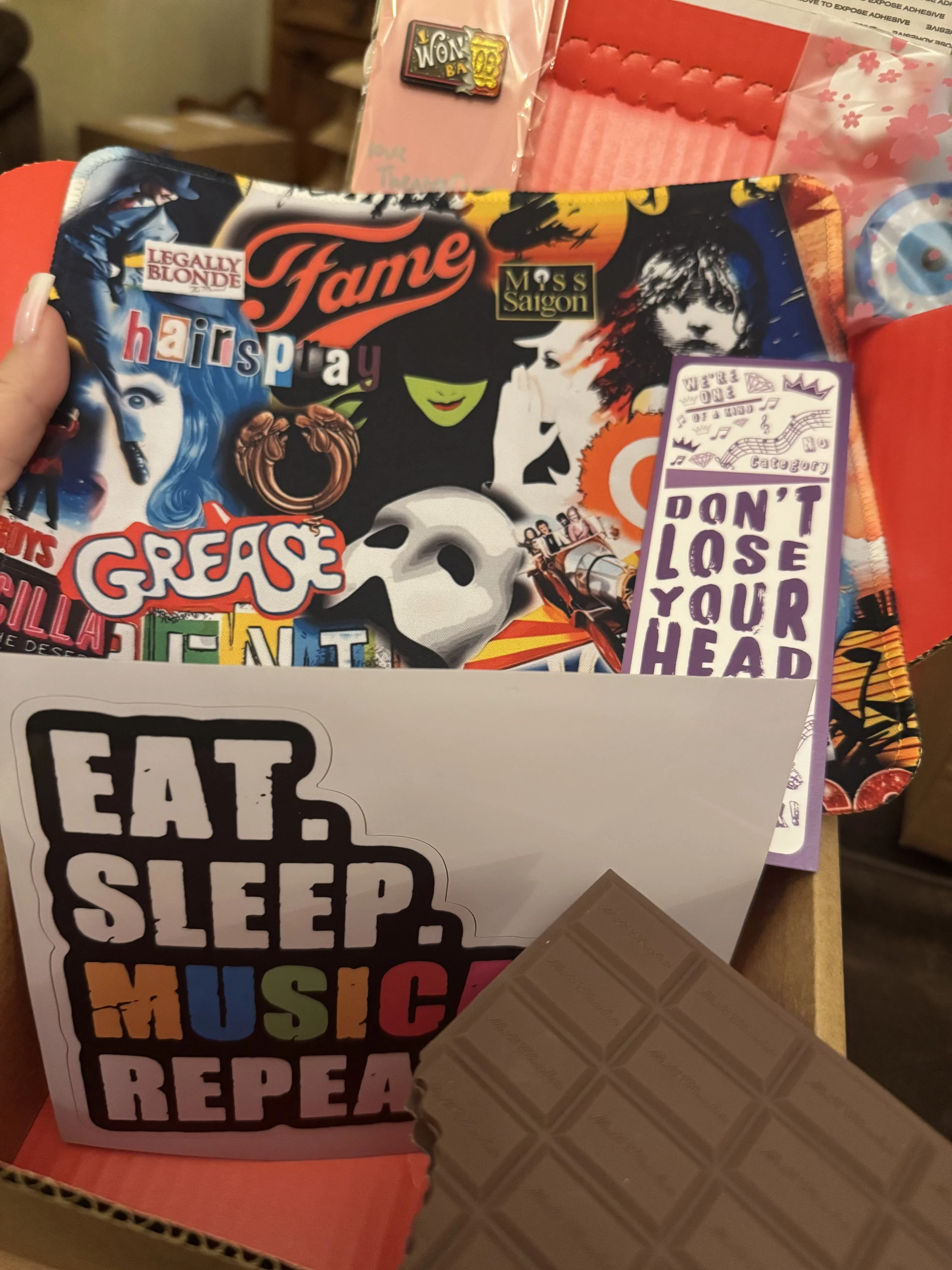 A box containing colorful stickers, a chocolate bar, and stationary items such as note cards with positive messages. The stickers include various designs and phrases like 'fame,' 'Grease,' and a cartoon face.