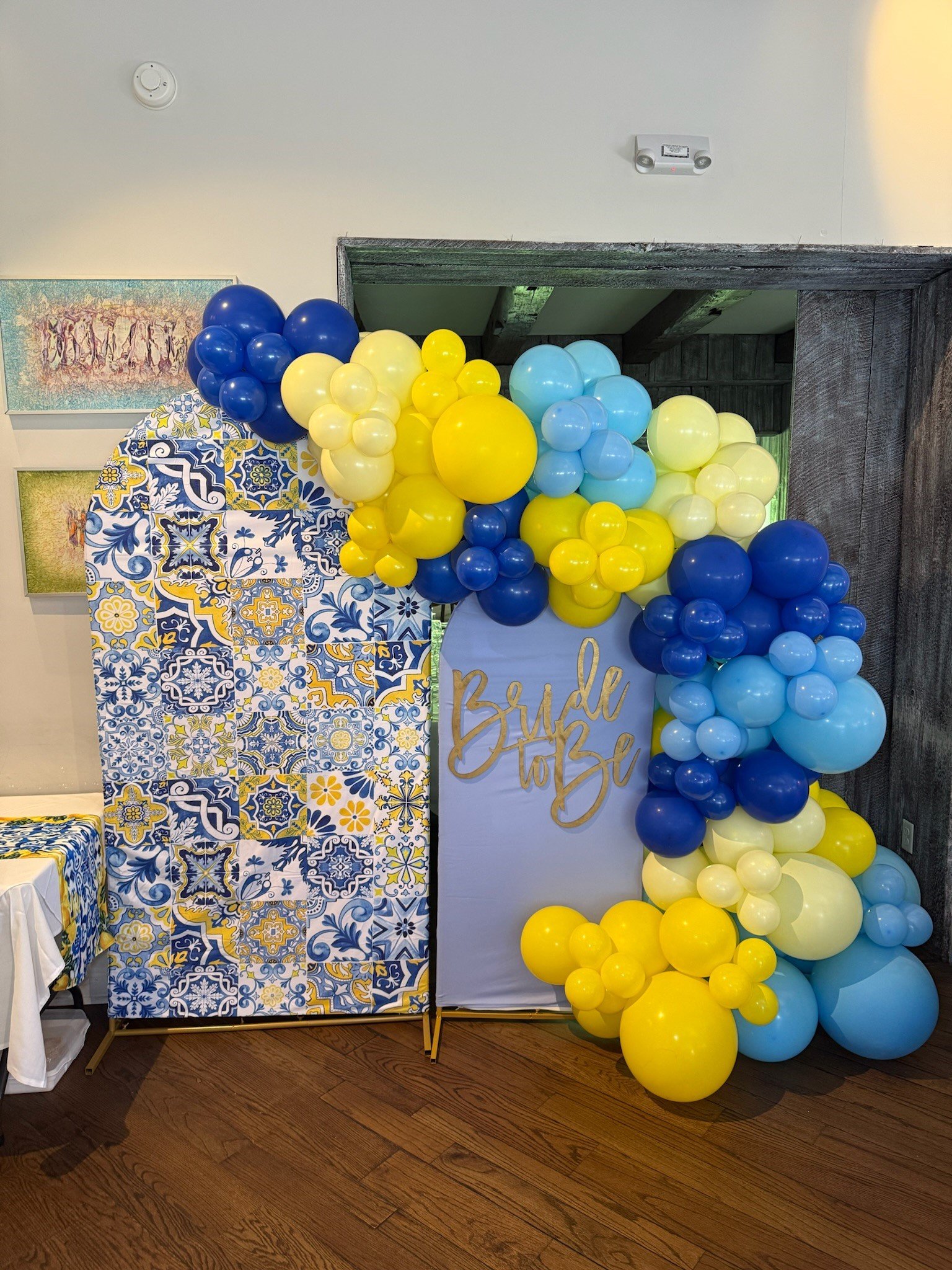 Party decoration with a balloon arch in shades of yellow, blue, and light green around a white sign that says 'Bride to Be.' Decorative tiles with floral and geometric patterns are on a standing board, and part of a table with a white tablecloth is visible on the left.