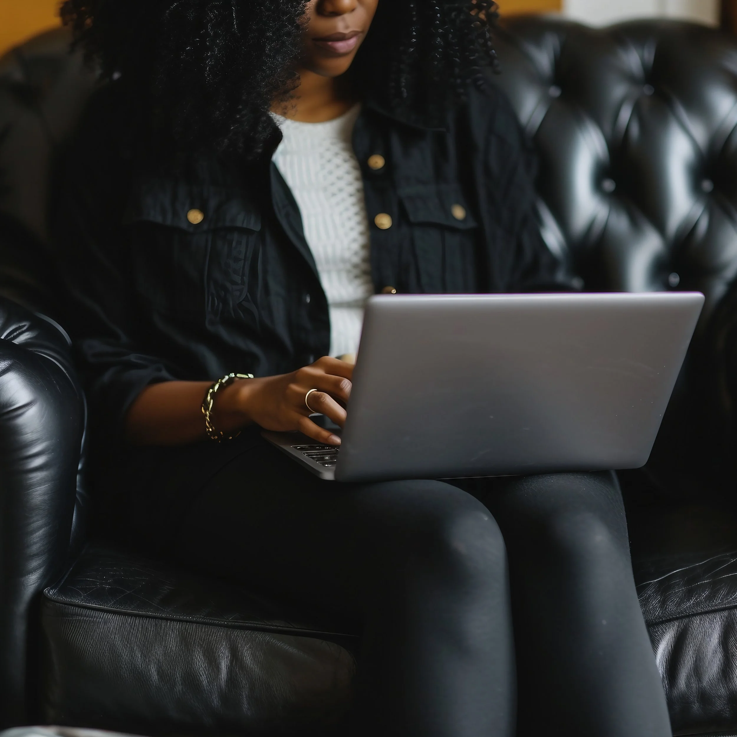 A woman sitting on a black leather couch using a silver laptop.