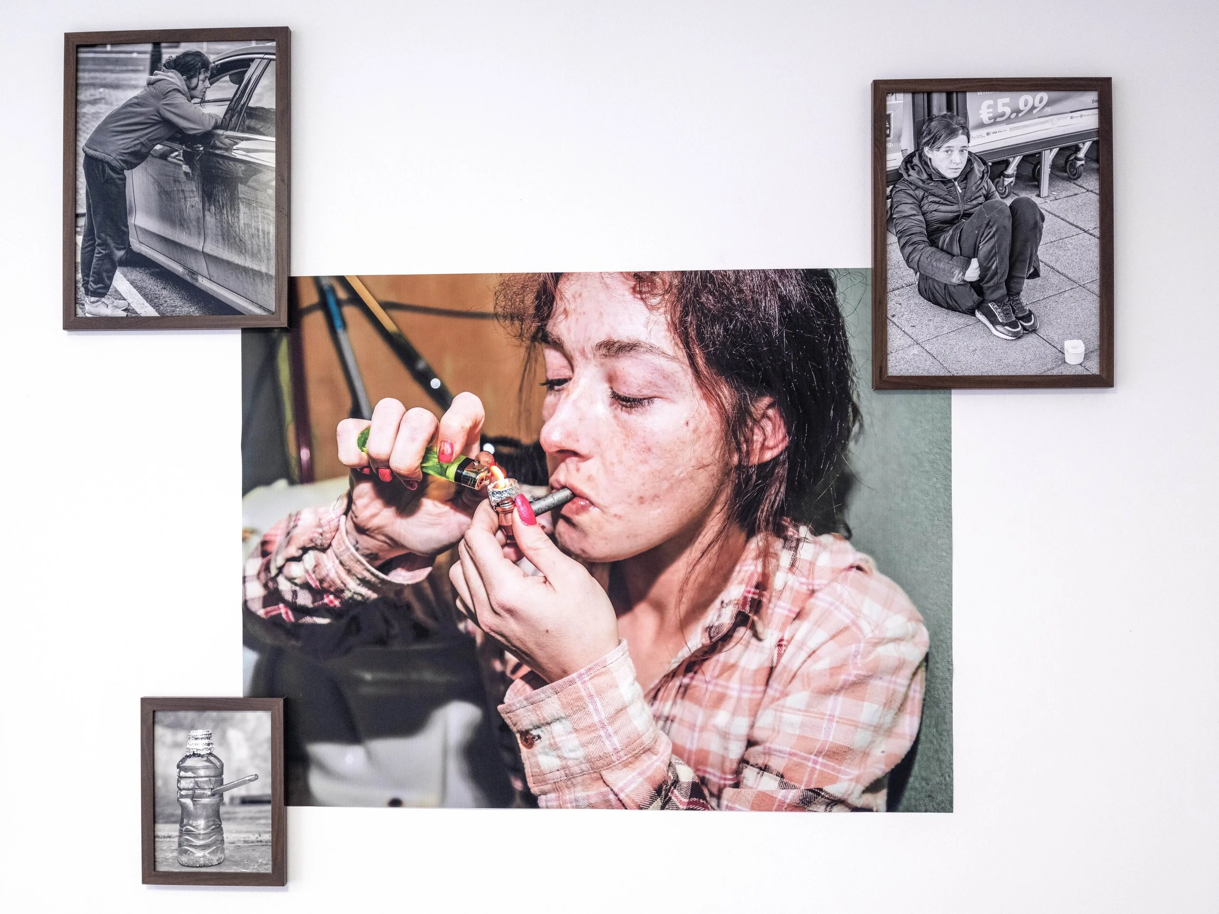 A young woman with dark curly hair smokes a joint, with her eyes closed and a relaxed expression, wearing a pink plaid shirt. Surrounding her are three framed photographs: one black-and-white photo of a boy leaning on a car, another black-and-white p