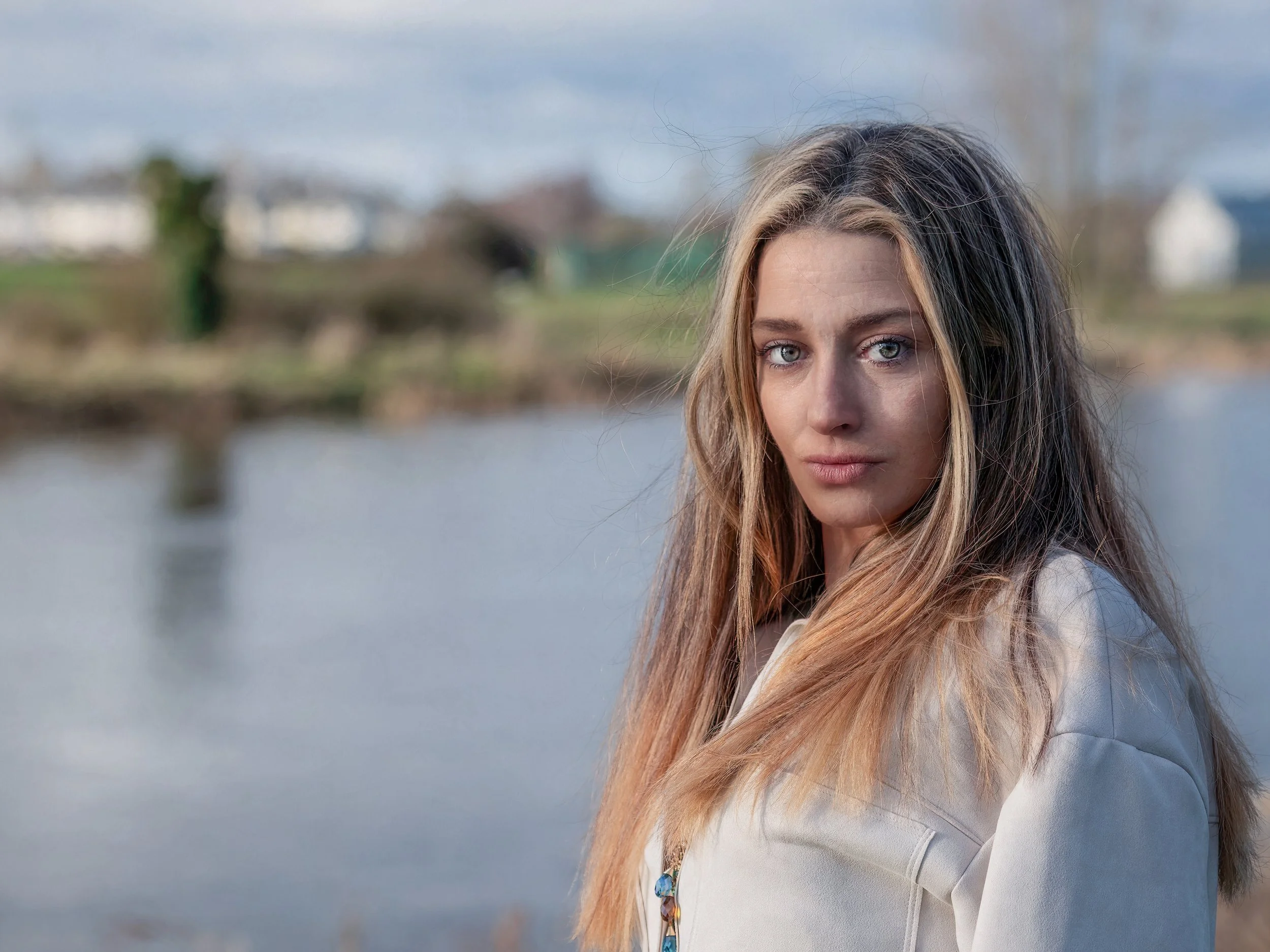 A woman named Antoinette Foley with long blonde hair and blue eyes standing outdoors near the River Barrow in Athy, Co. Kildare, Ireland with blurred houses in the background.