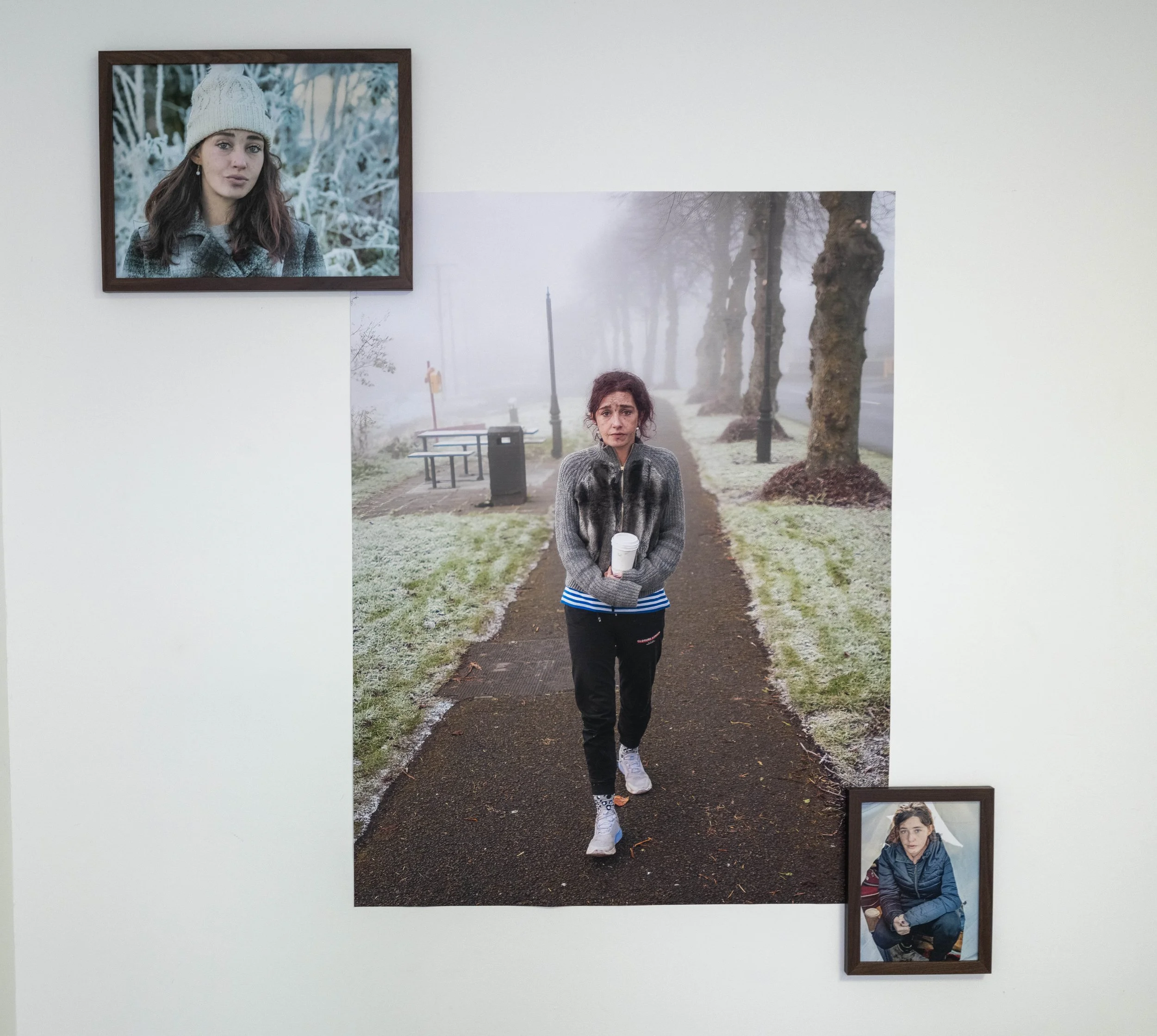 A woman walking on a foggy park pathway, holding a coffee cup, with two framed photographs of her on the wall behind her.