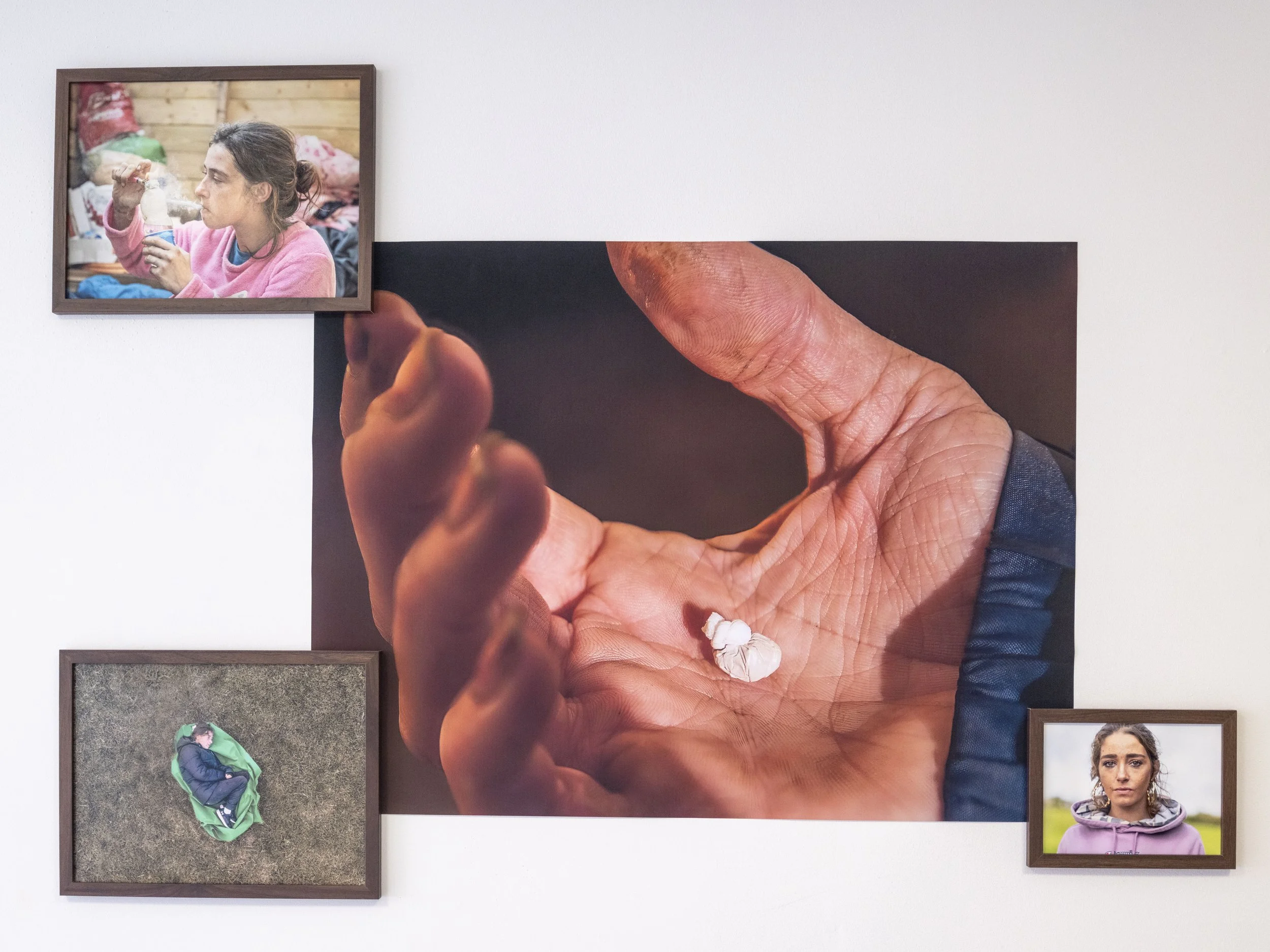 An art installation featuring a large photograph of a hand with a small white object, surrounded by three framed photographs: one of a woman eating, one of a person curled up on grass, and one of a woman outdoors.