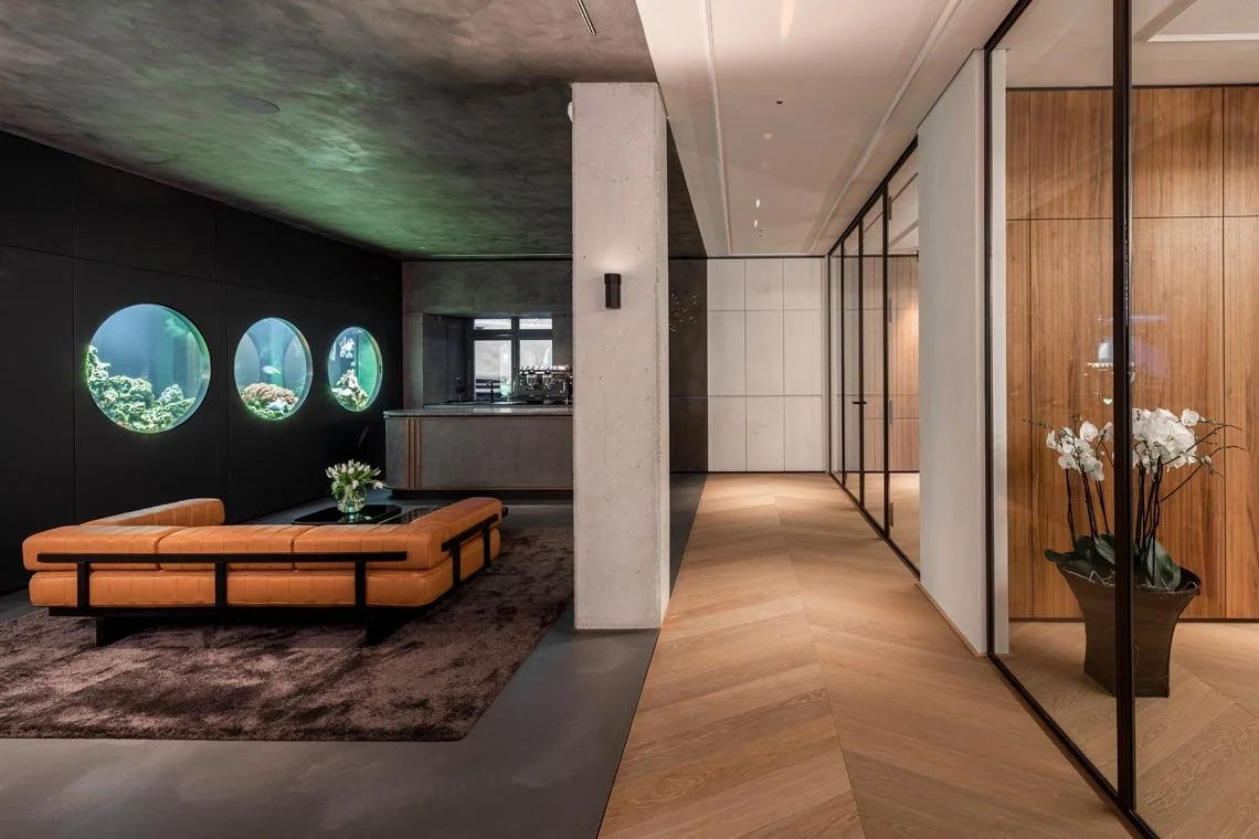 Modernes Lounge mit drei Aquarien, einem braunen Sofa, einem kleinen Beistelltisch mit Blumen, Holz- und Glaswänden.