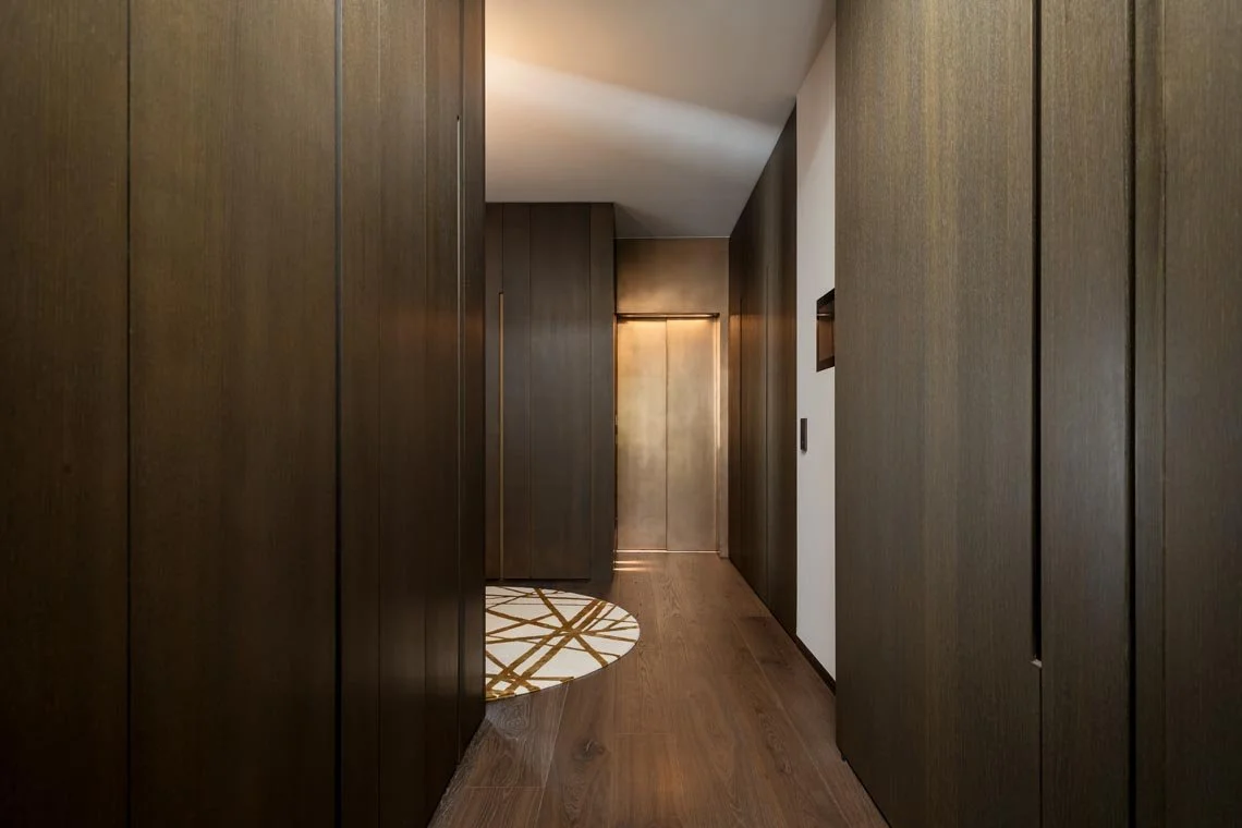 Ein Flur mit dunklen Holzschränken an den Wänden, einem runden Teppich mit geometrischem Muster und einer geschlossenen goldenen Tür im Hintergrund.