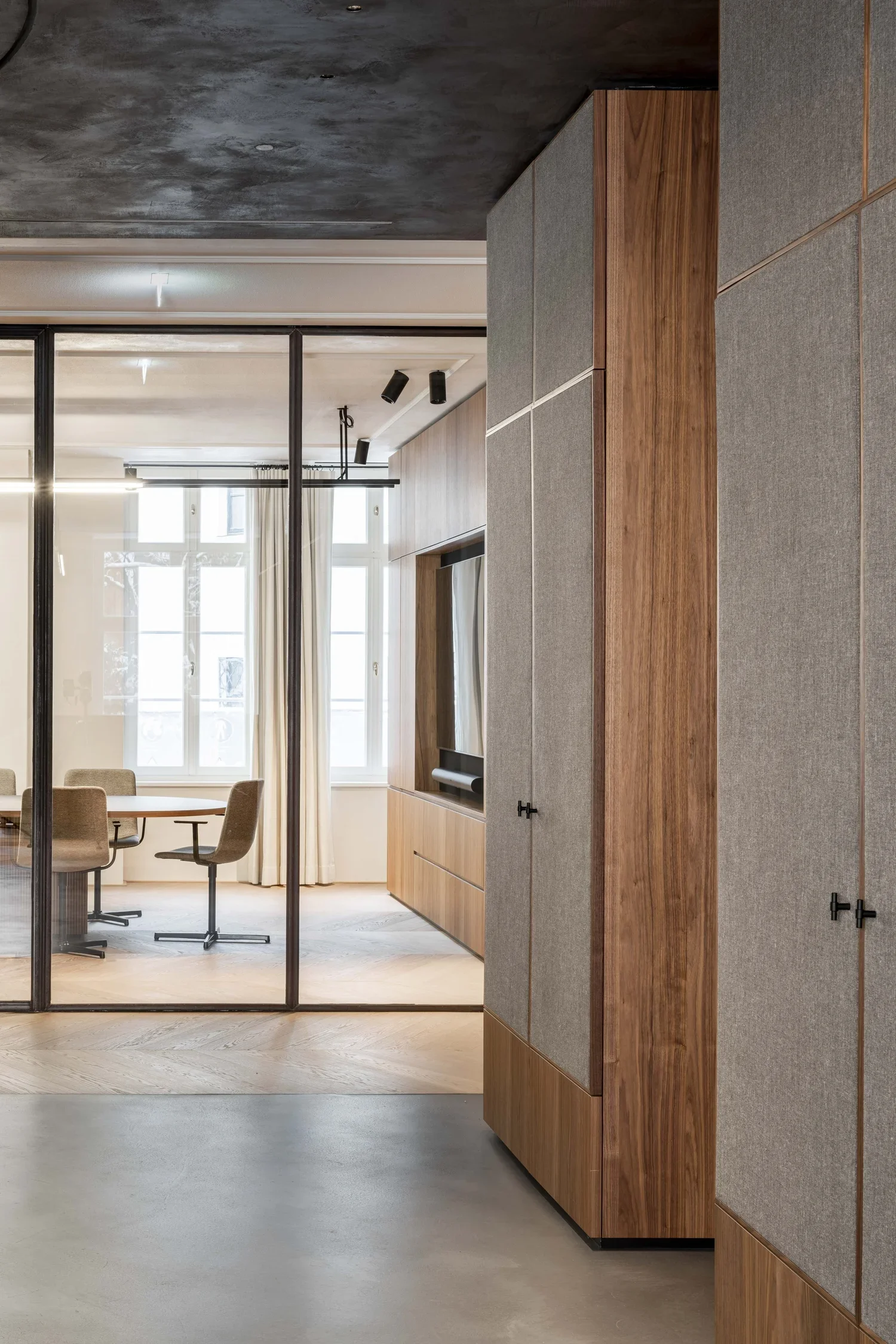 Innenraum eines modernen Büros mit Sicht auf einen Besprechungstisch und Stühle, durch eine Glaswand getrennt, mit Holz- und Stoffelementen in der Einrichtung.
