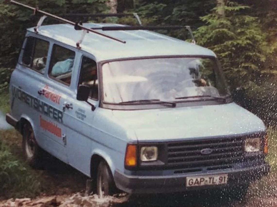 Ein blauer Van fährt durch eine Wasserpfütze auf einer Straße, umgeben von Bäumen, mit einem Hund im hinteren Fenster.