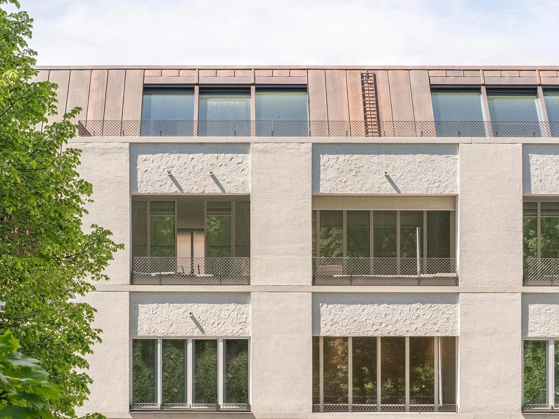 Ansicht eines modernen Mehrfamilienhauses mit drei Stockwerken, weißen Fassaden und großen Fenstern, mit Bäumen im Vordergrund.