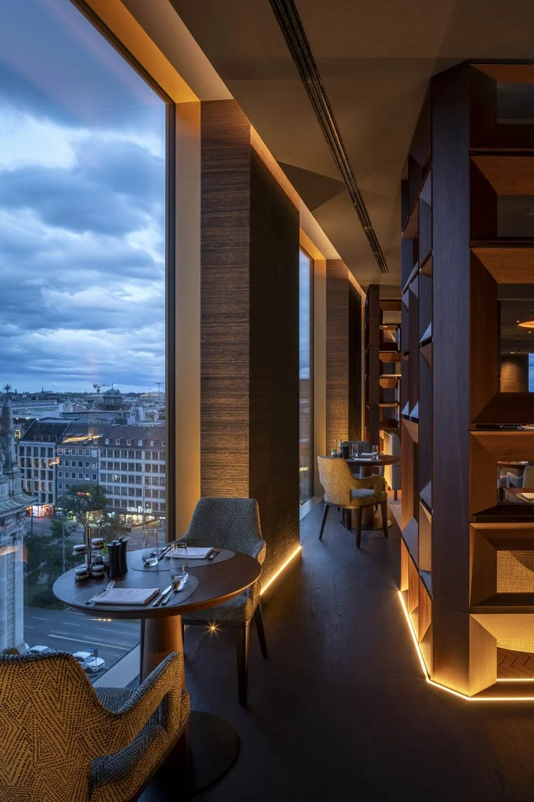 Innenaufnahme eines modernen, eleganten Restaurants mit großen Fenstern, die eine städtische Skyline bei Sonnenuntergang zeigen, und stilvoller Holzmöblierung.