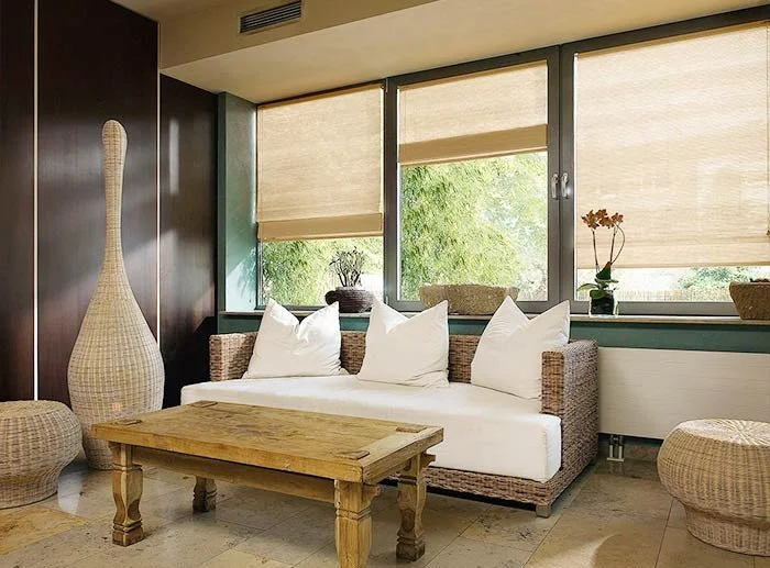 Innenraum eines Wohnzimmers mit einem weißen Sofa, einem Holzstuhl, großen Fenstern mit beige Jalousien und Dekorationsgegenständen wie einer Vase mit Blumen.