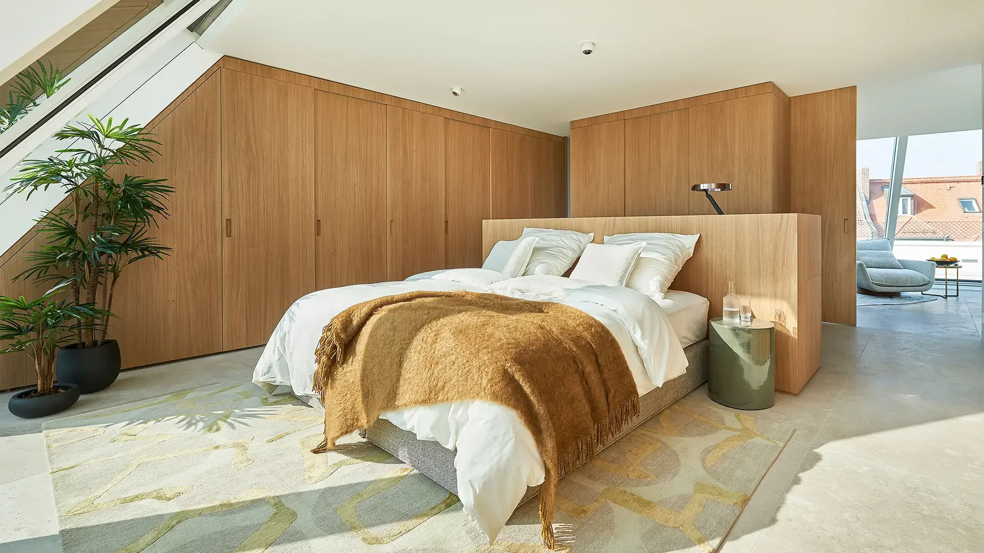Schlafzimmer mit Bett, weißen Bettwäsche und braunem Decke, Holzwand und Schränken, Pflanzen und Sitzbereich im Hintergrund, große Fenster mit Blick auf Dächer.