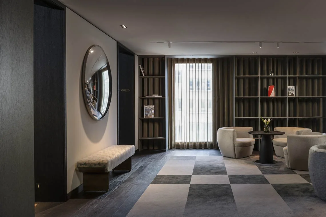 Moderner Hotel- oder Wohnraum mit beigen Sesseln, einem runden Tisch mit Vase, einem großen Fenster mit Vorhängen und Bücherregale an der Wand.