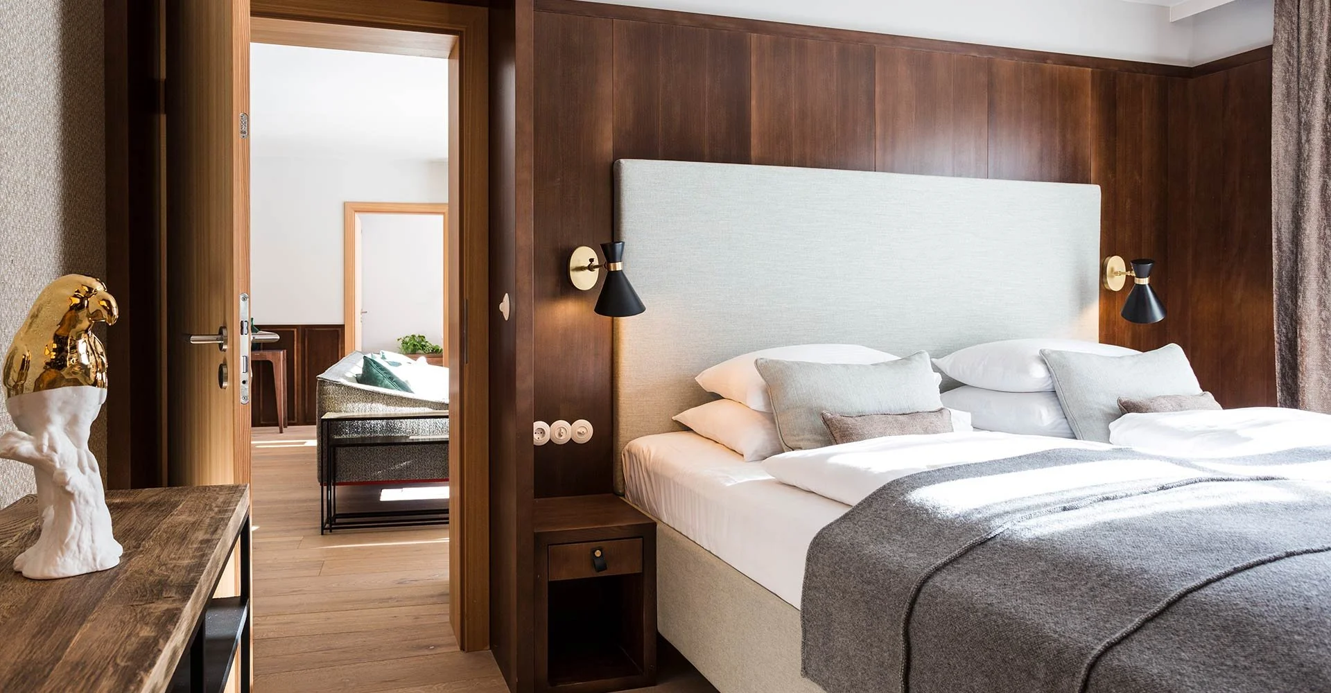 Ein Schlafzimmer mit Doppelbett, Holzwandverkleidung, Nachttischen und Wandlampen, Blick auf ein angrenzendes Zimmer mit Sitzmöglichkeit.