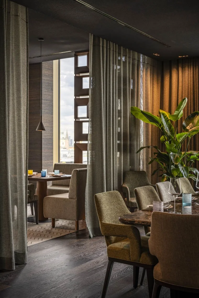 Ein elegantes Restaurant mit mehreren Tischen, Sitzgelegenheiten, einer großen Pflanze und Vorhängen, die einen Blick auf die Stadt im Hintergrund bieten.