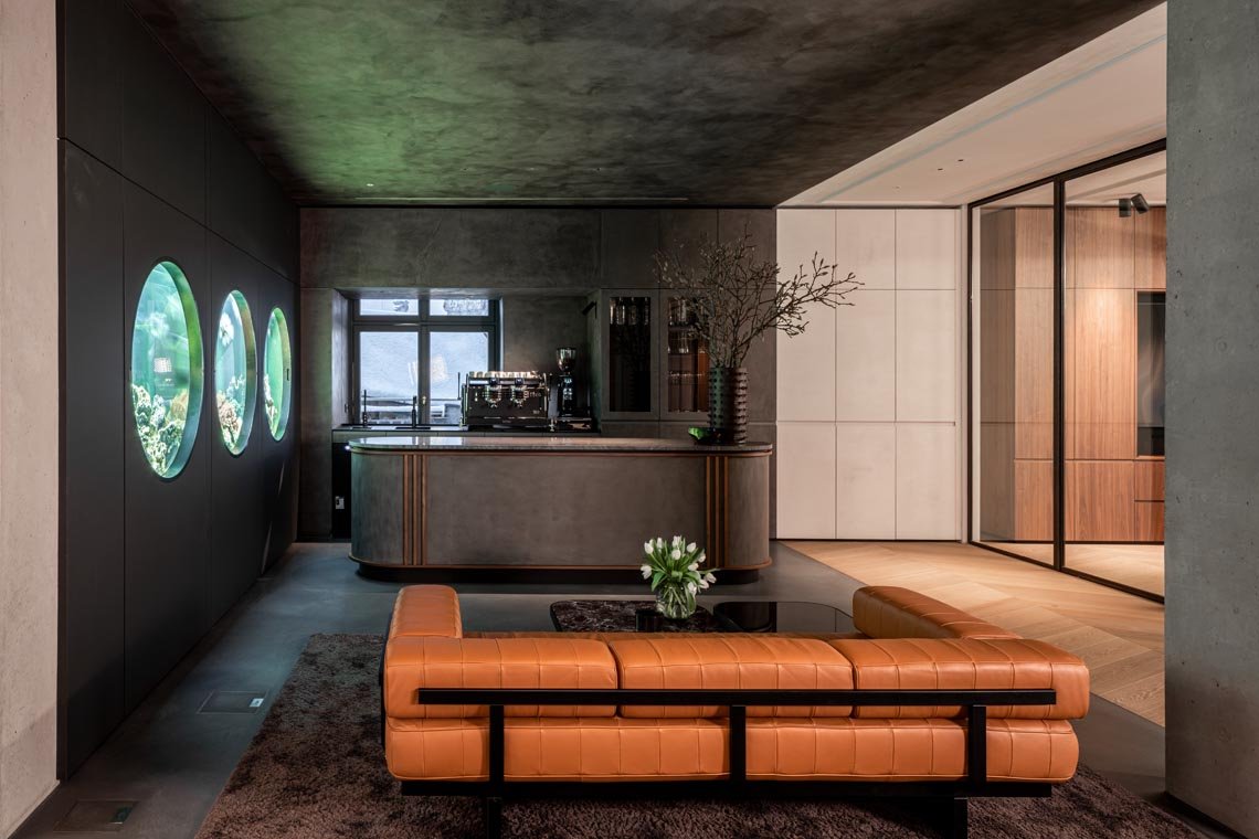 Moderne Wohnung mit grauem Betoninterieur, runden Aquariumfenstern an der linken Wand, braunem Leder-Couch, schwarzem Couchtisch mit Blumen und einer offenen Küche im Hintergrund.