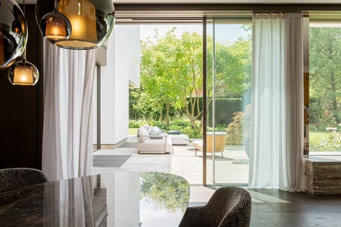 Innenraum mit Blick auf einen Garten, große Glasschiebefenster, weiße Vorhänge, dunkler Esstisch, moderne Beleuchtungskugeln.