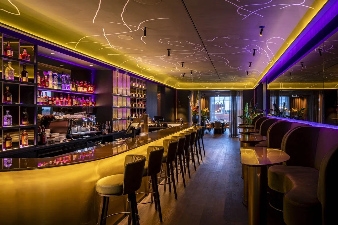 Luxusbar mit dunklem Interieur, goldenem Tresen und leuchtenden Regalen mit Spirituosen, bequemen Sitzen und indirekter Beleuchtung.