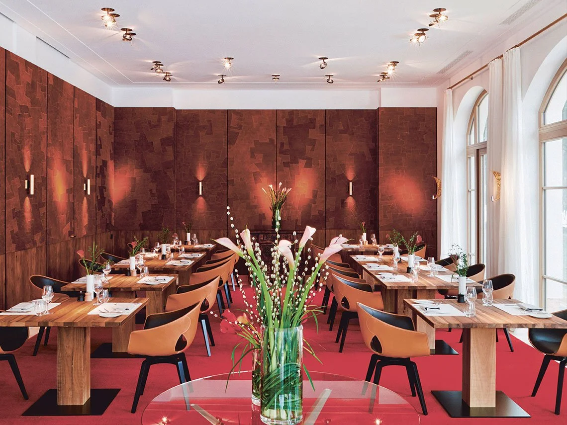 Ein eleganter Speiseraum mit Holztischen, schwarzen und beigen Stühlen, weißen Tischdecken und Gläsern, große Fenster mit weißen Vorhängen, rote Teppichboden, zentrale Blumenvasen mit weißen Blumen, moderne Wandlampen und eine rote Wand im Hintergrun