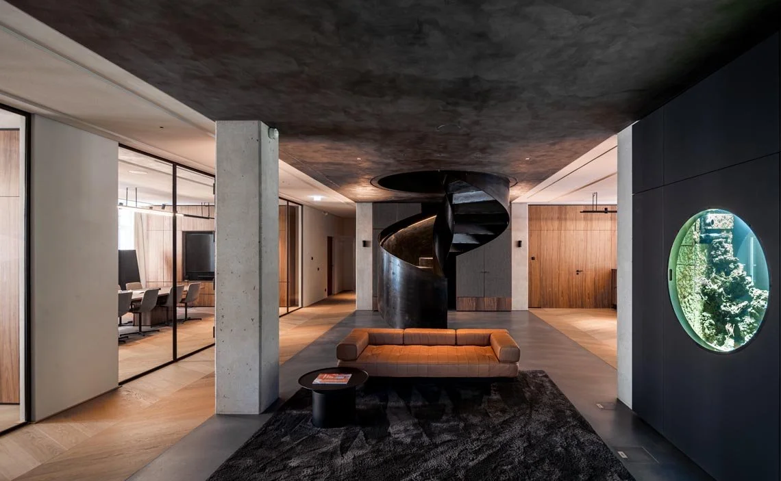 Innenansicht eines modernen Wohnbereichs mit schwarzer Spiraltreppe, Sofa, ovalem Aquarium und Holz- und Betonelementen.