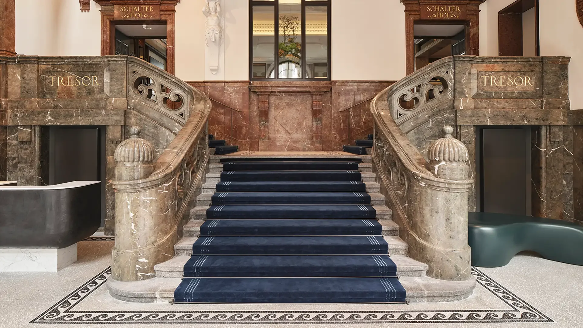 Elegante Treppe im Hotel mit schwarzem Teppich, braune Marmorskulpturen, goldene Beschriftungen 'Schalter Hof' und 'Tresor' an den Wänden, hohe Decke mit Kandelaber.