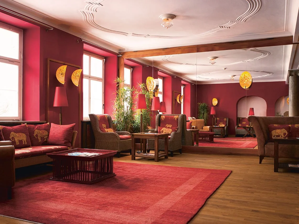 Eleganter, gemütlicher Lounge-Bereich mit roten Wänden, samtigem Sitzmöbel mit Tiermustern, Pflanzen, roten Lampen und weichem Teppich in warmen Tönen.