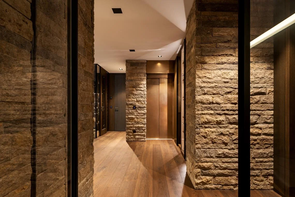 Innenansicht eines modernen Flurs mit Holz- und Steinelementen, beleuchtet mit Decken- und Wandlicht, führt zu einem Aufzug.