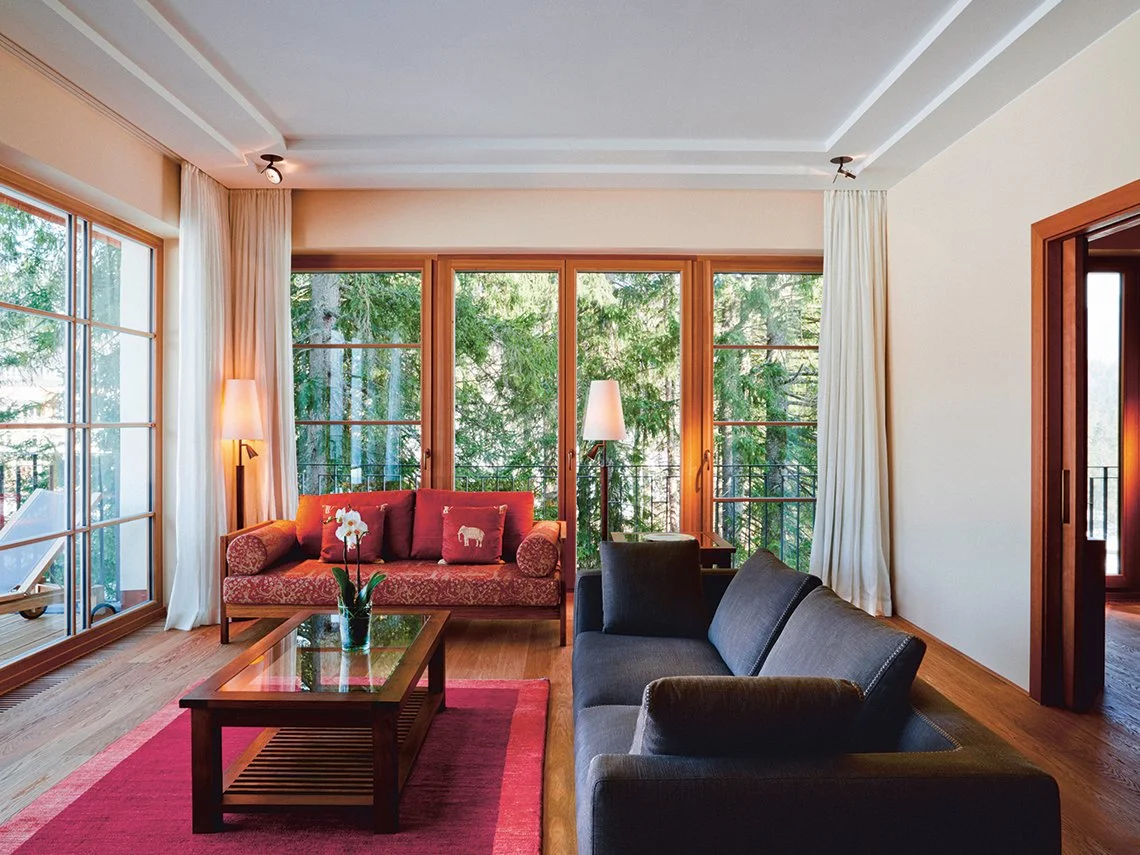 Wohnzimmer mit großen Fenstern, Holzmöbeln, grauem und rotem Sofa, Teppich und Pflanzen, umgeben von Bäumen im Freien.