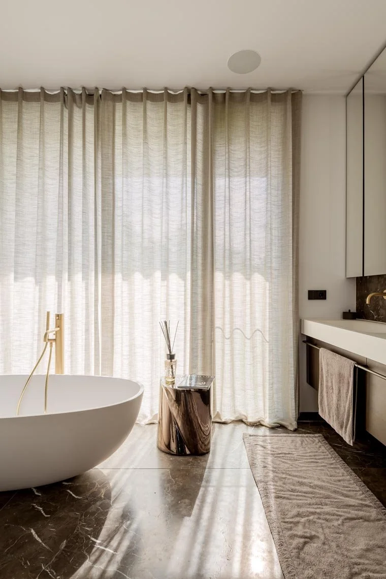 Ein modernes Badezimmer mit einer weißen Wanne, einem kleinen Beistelltisch aus Holz, langen weißen Vorhängen, einem Waschbecken mit Handtuch und grauem Teppich auf einem schwarzen Fliesenboden.