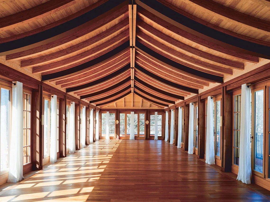 Ein großer, gut erleuchteter Raum mit einer gewölbten Holzbalkendecke, Holzboden und großen Fenstern mit weißen Vorhängen, die Sonnenlicht hereinlassen.