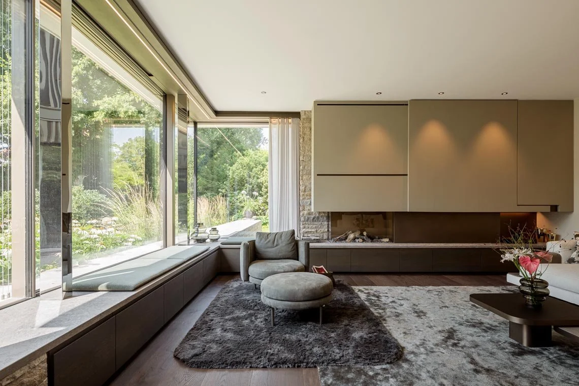 Moderne, helle Wohnzimmer mit großen Fenstern, gemütlicher Sitzcouch, Sessel mit Hocker, Kamin, schöne Dekorationen und Blick ins Grüne