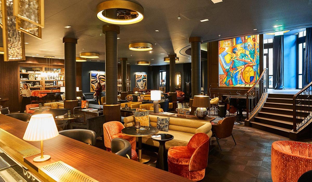 Elegantes Lounge mit gemütlichen Sitzbereichen, bunten Sesseln, Stehlampen, abstrakter Kunst an den Wänden und großen Fenstern mit blauen Vorhängen.