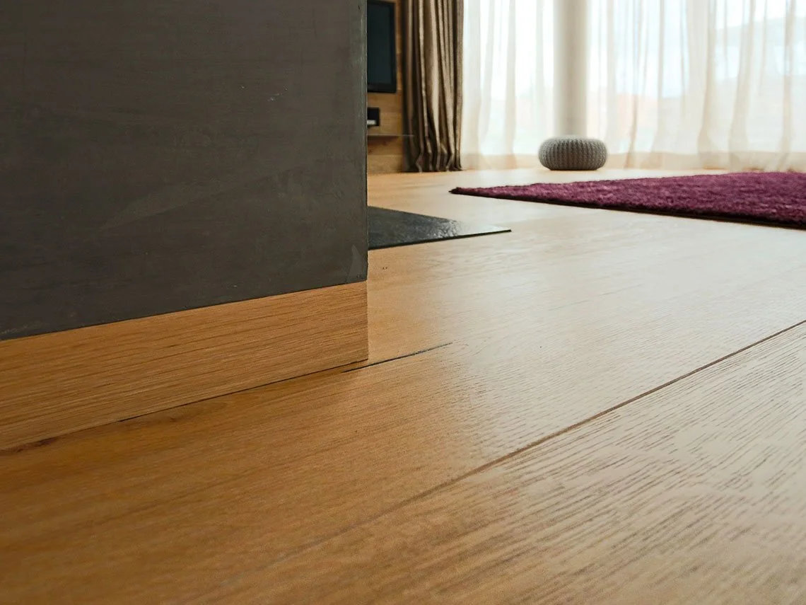 Innenaufnahme eines Wohnzimmers mit Holzboden, Vorhang und Teppich, Blick auf einen Teil des Zimmers mit einem schwarzen Möbelstück, einem runden Kissen und einem Fernseher im Hintergrund.