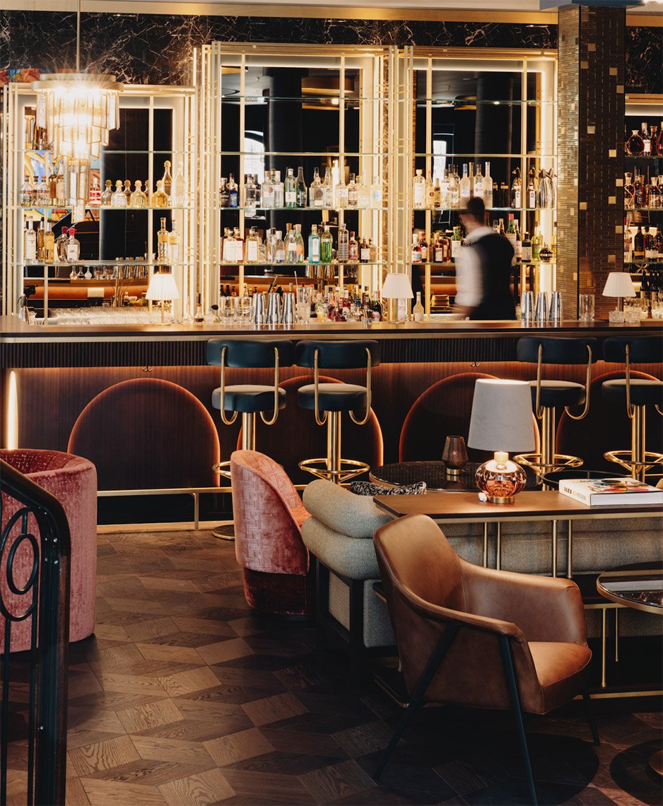 Ein eleganter Bar- und Lounge-Bereich mit dunklen Holzboden, bequemen Sesseln und einem kunstvoll gestalteten Barregal mit Flaschen und Schnäpsen im Hintergrund. Helligkeit durch Lampen und ein großes Kronleuchter